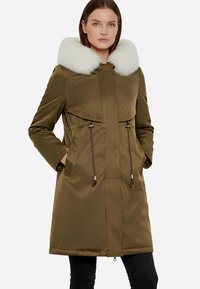 Parka verde oliva con cappuccio in pelliccia sintetica bianca, chiusura con zip, vita regolabile con cordoncino e due tasche frontali. Tessuto lucido e compatto con una texture liscia.