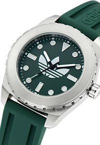 Montre ronde avec un cadran vert mat, des index argentés et trois aiguilles. Boîtier en acier inoxydable avec une lunette cannelée et un bracelet en caoutchouc vert.