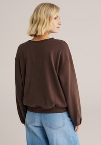 Rudas medvilninis džemperis su laisvu siluetu, nusvirusiais pečiais ir ribiniu apvadu, dėvimas su šviesiai mėlynomis, aukšto liemens džinsais.