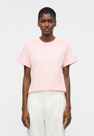 Γυναίκα με σκουρόχρωμο δέρμα και κοντά μαλλιά, ντυμένη με ανοιχτό ροζ κοντομάνικο πουκάμισο και λευκό παντελόνι κοψίματος, στέκεται μπροστά σε απλό φόντο.