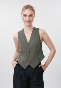 Jeune femme aux cheveux blonds portant un gilet gris sans manches boutonné et un pantalon noir, debout les mains dans les poches.