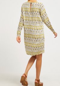 Robe à manches longues avec un motif géométrique jaune et gris, fente dans le dos et coupe ample. Portée avec des sandales compensées marron.