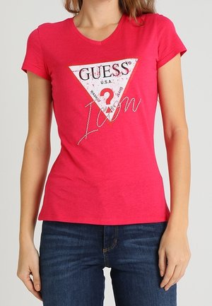 Różowa koszulka z krótkim rękawem i dekoltem w serek, z trójkątną białą grafiką, na której widnieje napis "GUESS? U.S.A." oraz "I am" w cyrkoniach.