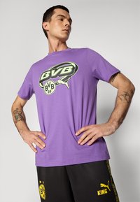 Mand iført en lilla BVB-logo t-shirt og sorte shorts med gule BVB- og Puma King-logoer, stående med hænderne i siden.