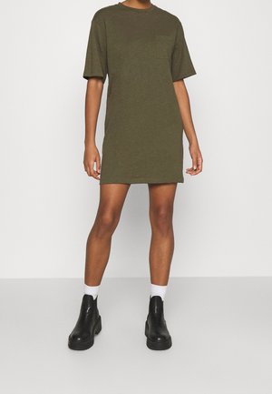 Osoba ubrana w oliwkową sukienkę z krótkim rękawem i kieszonką na piersi, do tego białe skarpetki i czarne masywne botki, stojąca na szarej podłodze.