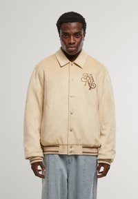 Bež jakna od umjetnog zamka s ovratnikom, logotipom na prsima, dugmadi na kopčanje i rebrastim manžetama i rubom. Uparena s svijetloplavim trapericama.