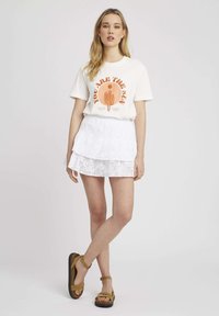 Jupe blanche à volants avec des détails en œillets, t-shirt crème avec un design graphique orange, et sandales à lanières brunes.