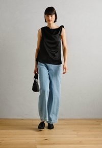 Top de satén negro sin mangas con un diseño drapeado y detalle de un solo hombro. Jeans de mezclilla de pierna ancha en azul claro. Bolso y zapatos negros.