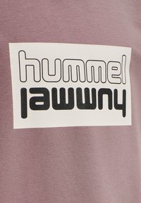Camiseta de algodón lavanda con un gran gráfico rectangular crema que presenta un texto en negrita negro que dice "hummel jawwry". Textura suave.