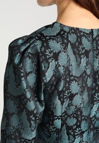 Černý látkový top s propracovaným teal květinovým vzorem, nařasenými rukávy a nenápadným zapínáním na zip na zadní straně. Hladká textura.