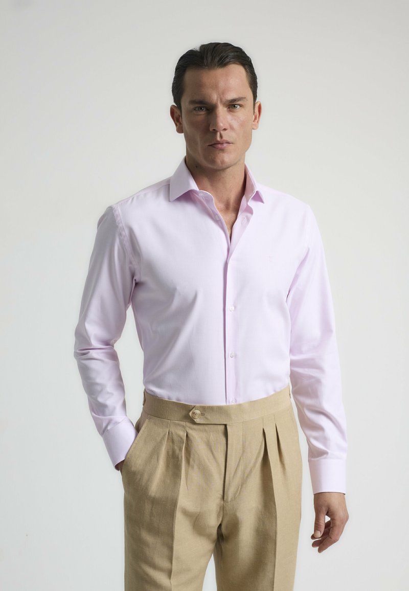 Camisa de manga larga de color rosa claro con cuello abotonado, combinada con pantalones beige de corte a medida con pliegues y detalle de botón.