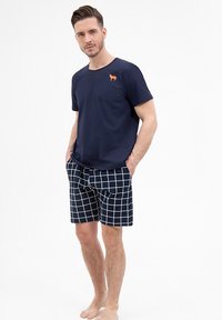 Granatowa koszulka z małym pomarańczowym logo psa, połączona z granatowymi szortami z białym wzorem w kratkę. Szorty mają elastyczny pas.