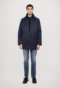 Navy blue, knee-length coat with a padded texture, button closure, and side pockets. Worn over a black sweater, paired with blue jeans and black shoes.