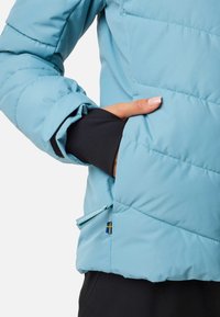 Personne portant une veste matelassée bleu clair, la main glissée dans la poche latérale, montrant une extension de manche intérieure noire et un onglet partiel de la tirette de la fermeture éclair.
