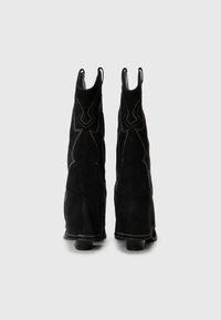 Bottines en suede noir avec une forme évasée, présentant des coutures blanches contrastantes et des languettes à l'arrière pour un enfilage facile.