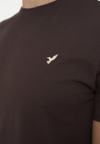 Primo piano di una maglietta marrone con un piccolo ricamo bianco a forma di uccello sul lato sinistro del petto.