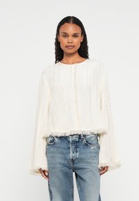 Frau mit lockigem Haar, die ein lockeres, weißes, strukturiertes Langarmtop mit ausgefransten Kanten und blaue Jeans trägt, vor einem schlichten Hintergrund.