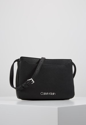Sac bandoulière rectangulaire noir avec sangle réglable et logo Calvin Klein argenté à l'avant, présenté sur un piédestal blanc.