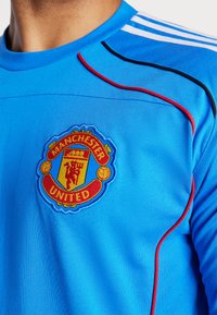 Mėlyna sportinė marškinė su trumpomis rankovėmis, su raudonais ir juodais šonais bei išsiuvinėta Mančesterio „United“ emblema ant krūtinės.