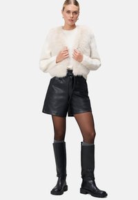 Faux-Fur-Jacke in Creme mit breitem Kragen; schwarze Leder-Shorts mit gebundenem Bund; knielange schwarze Stiefel; transparente Strumpfhose mit grauen Socken.