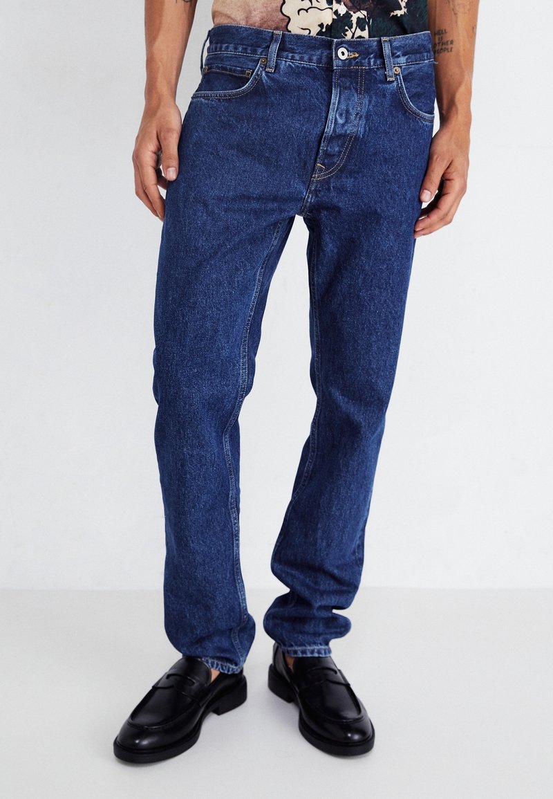 Hombre vestido con jeans de mezclilla oscura y mocasines de cuero negros, de pie contra un fondo blanco liso.