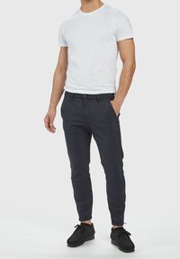 Pantalons slim-fit navy, tissu texturé, jambe fuselée, poches avant ; portés avec un t-shirt blanc ajusté et des chaussures décontractées noires.