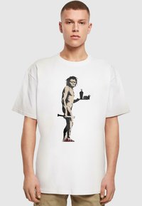 Tricou alb din bumbac cu un desen imprimat care prezintă o figură ținând un os și un tavă de fast-food, reprezentând un om al peșterii într-un stil umoristic.