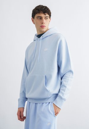 Fiatal férfi világoskék Nike kapucnis pulóverben és hozzá illő melegítőnadrágban, balra néz, kezét zsebében tartja, egyszínű fehér háttér előtt.