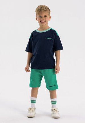 T-shirt marine avec des accents teal et le texte "AMUSEMENT ILLIMITÉ", associé à un short vert. Chaussettes blanches avec des rayures vertes et baskets blanches.