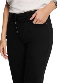 Jeans de mezclilla negros de talle alto con cierre de botones, que cuentan con cinco bolsillos y una textura suave, llevados por una persona con una mano en el bolsillo.