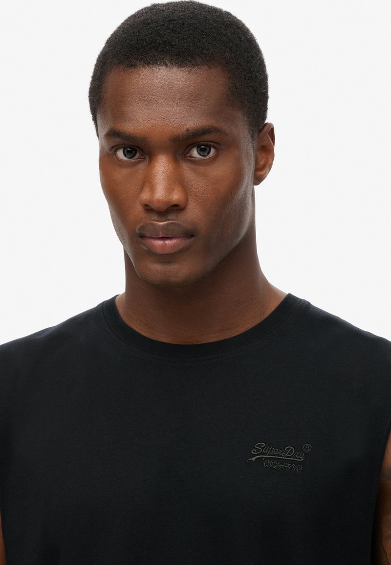 Camiseta negra sin mangas con un cuello redondo. Presenta un discreto logo bordado en el lado izquierdo del pecho. Textura de algodón suave.