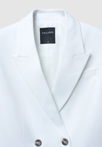 Blazer bianco doppiopetto realizzato in tessuto liscio, con colletto a punte, dettagli di tasche sottili e bottoni metallici. Etichetta del marchio visibile.