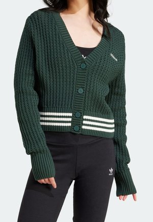 Femme portant un cardigan en maille vert foncé avec des rayures blanches et des boutons, associé à des leggings noirs Adidas.