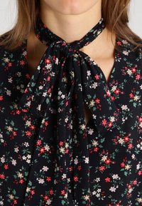Blusa nera a fiori con un colletto a fiocco. Piccole fiori rossi, rosa e bianchi sono sparsi su tutta la superficie, accentuati da foglie verdi.