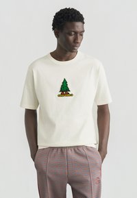 Μπεζ βαμβακερό μπλουζάκι με μπαλώμα σε σχήμα πράσινου δέντρου και κείμενο «Converse» από κάτω. Συνδυασμένο με καρό παντελόνια σε κόκκινο και μπλε.