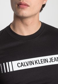 Mladý člověk v černém tričku s kulatým výstřihem, které má bílý horizontální pruh a text "Calvin Klein Jeans" přes hrudník.
