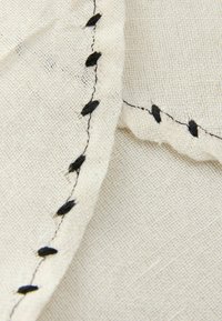 Off-white stof met een textuurafwerking, voorzien van zwarte stitching en decoratieve accenten langs de rand in een consistent patroon.