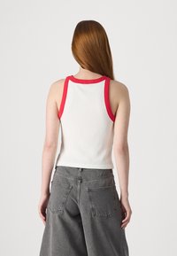 Débardeur blanc à côtes avec bordure rouge au niveau du col et des emmanchures, porté avec un jean large gris taille haute. Détails de couture sur le pantalon.