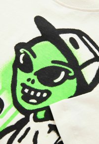 Bomuld t-shirt med et lyst grønt rumvæsen grafisk motiv med en sort kasket og kraftig sort kant, på en cremet baggrund med teksturerede printdetaljer.