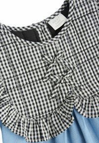 Musta-valkoinen ruudullinen mekko sinisellä hameella, jossa on röyhelökaula-aukko ja koristeelliset nauhat. Kangas näyttää sileältä. Brändilogo on näkyvissä.