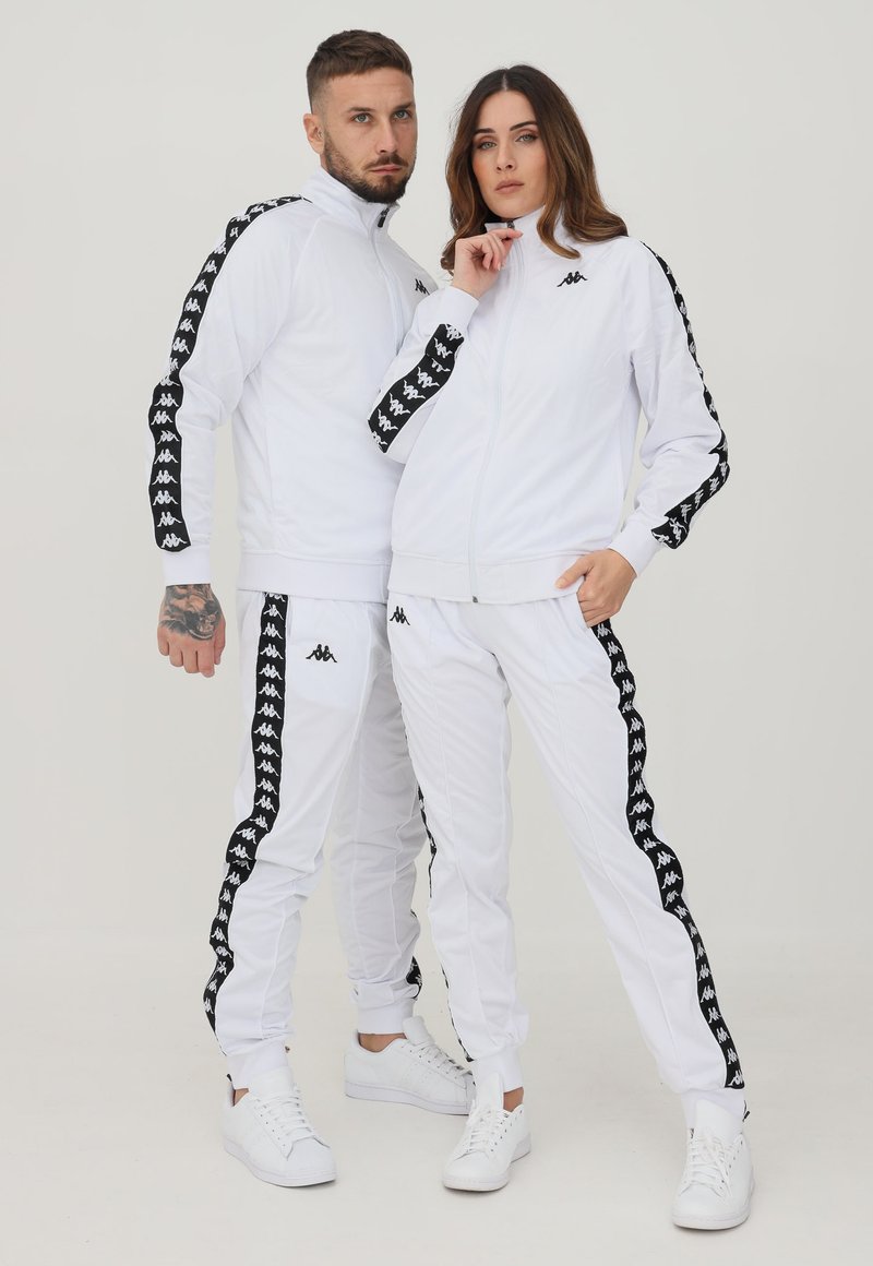 Uomo e donna che indossano tute bianche abbinate con strisce nere con logo sulle maniche e sulle gambe dei pantaloni, posando davanti a uno sfondo semplice.