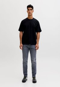 Ung mand står foran hvid baggrund iført sort oversized t-shirt, falmede grå jeans og sorte sneakers med neon grønne snørebånd.