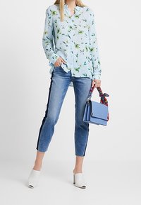 Ljusblå blommig skjorta med knappar, blå denim caprijeans med sidostråk, vita klackskor med blockklack och en liten ljusblå handväska med ett mönstrat band.