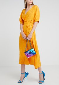 Vestido envolvente amarillo con mangas cortas abullonadas y textura floral; combinado con un bolso de mano metálico multicolor y sandalias de tacón azul.