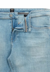 Jeans de denim azul claro con la cremallera parcialmente desabrochada, botón abrochado y etiquetas de la marca visibles en la cinturilla.