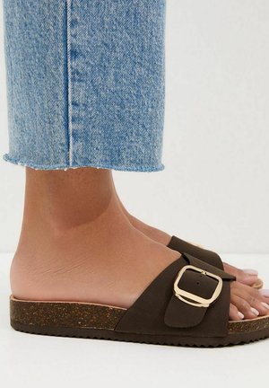 Sandales slides marron avec un détail boucle, semelle intérieure plate en liège et semelle texturée. Portées avec un jean denim bleu clair retroussé.