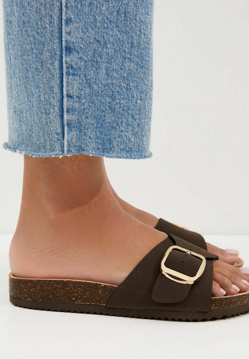 Brązowe sandały na płaskiej podeszwie z korka, z klamrą i teksturowaną podeszwą. Połączone z podwiniętymi jasnoniebieskimi jeansami.