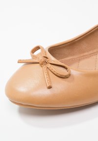 Ballerine beige clair en cuir lisse. Elle présente un bout arrondi et un petit nœud décoratif à l'avant, avec une semelle intérieure texturée.