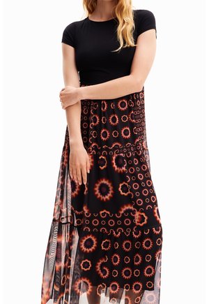 Žena nosící černý top s krátkým rukávem a černou sukni s oranžovými kruhovými květinovými vzory, držící jednu ruku přes tělo.