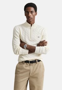 Cremefarvet kabelstrikket sweater med halvlange lynlåslukning ved kraven. Har ribbestrikkede manchetter og kant. Sammen med beige chinos og sort bælte. Sølvur synligt.